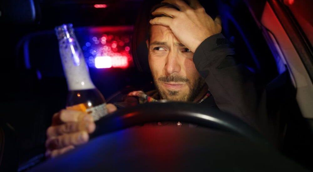 A person sits in a car holding a bottle, looking distressed. Police lights flash red and blue through the rear window, casting colored glows inside the vehicle.
