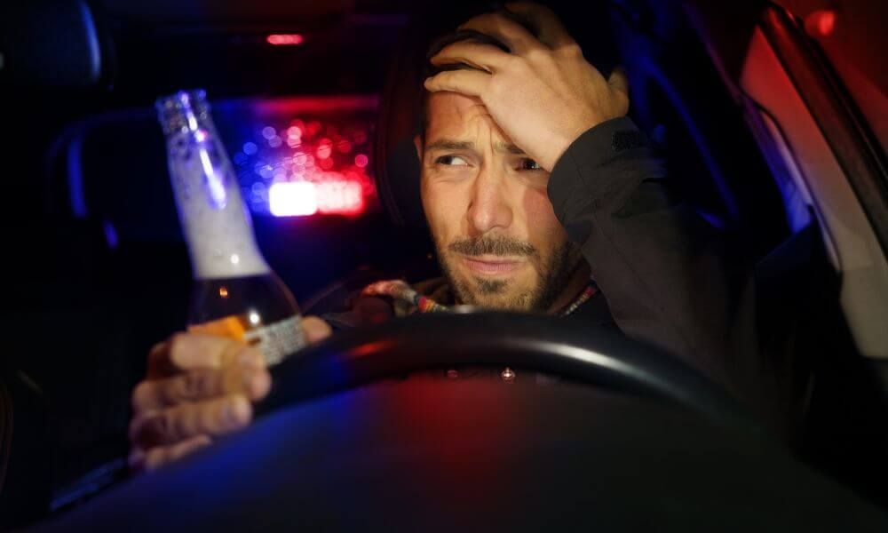 A person sits in a car holding a bottle, looking distressed. Police lights flash red and blue through the rear window, casting colored glows inside the vehicle.