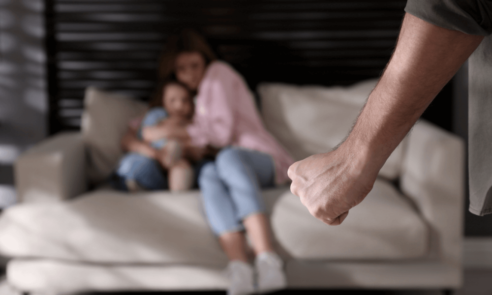 A clenched fist is prominently foregrounded, poised and tense. In the blurred background, a woman and child sit on a couch, appearing vulnerable in a room with soft lighting.