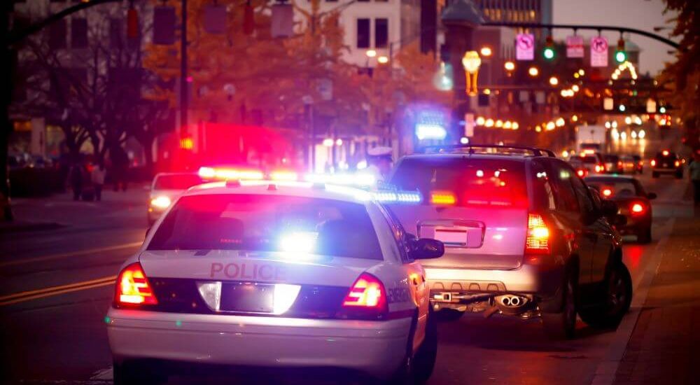 A police car, with flashing lights, is parked behind a pulled-over vehicle on a city street. Streetlights illuminate autumn trees and buildings as cars pass by in the background.