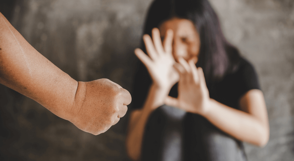A clenched fist is directed toward a woman shielding her face with her hands, set against a blurred gray background, suggesting a tense, confrontational scene.