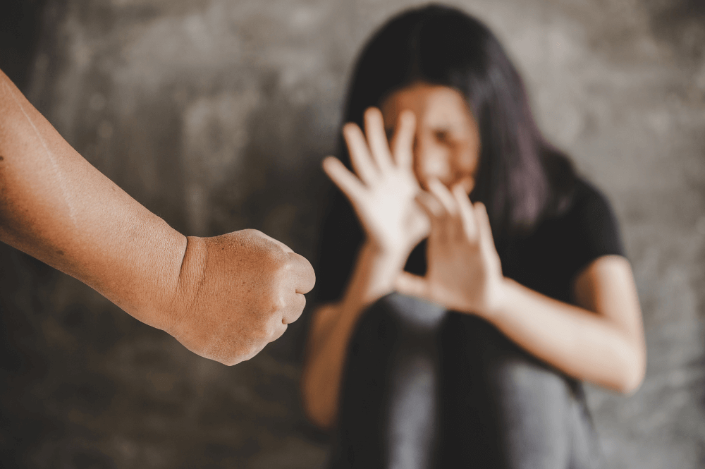 A clenched fist is directed toward a woman shielding her face with her hands, set against a blurred gray background, suggesting a tense, confrontational scene.