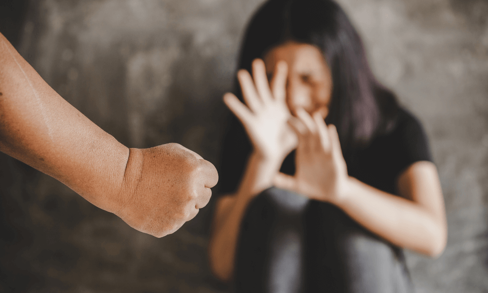 A clenched fist is directed toward a woman shielding her face with her hands, set against a blurred gray background, suggesting a tense, confrontational scene.