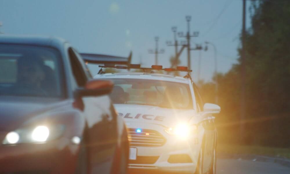 A police car with flashing lights is positioned on a road, interacting with a nearby red sedan. The environment appears dim, with power lines visible in the background.