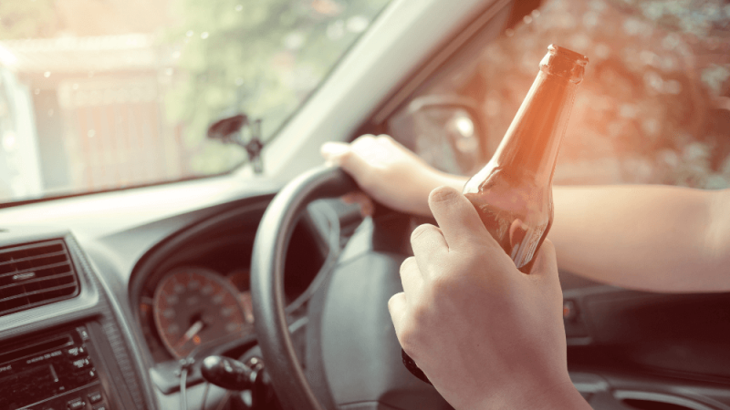 A hand holds a beer bottle while gripping the steering wheel of a car, with blurred trees and a fence visible through the windshield, suggesting a relaxed driving environment.