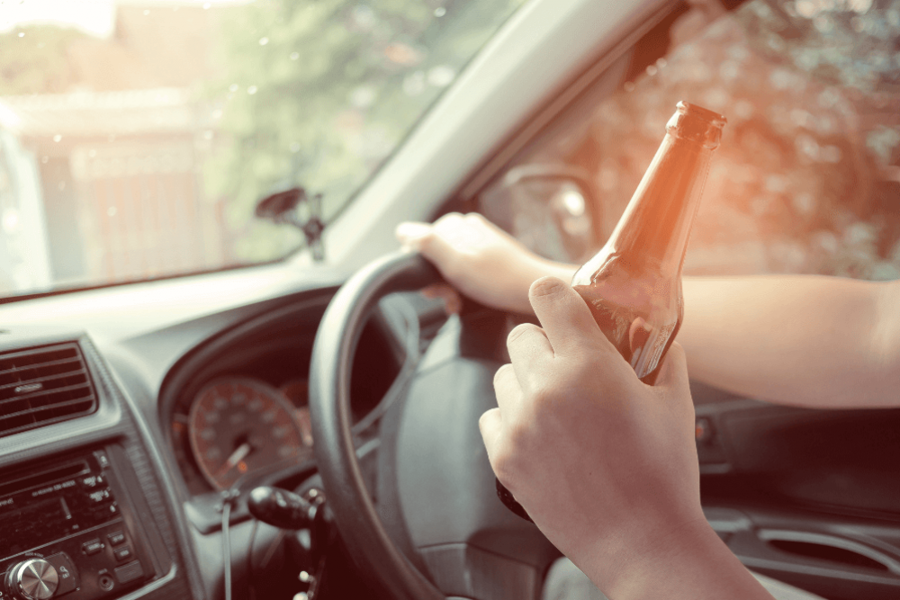 A hand holds a beer bottle while gripping the steering wheel of a car, with blurred trees and a fence visible through the windshield, suggesting a relaxed driving environment.