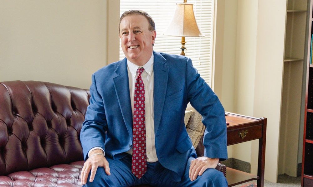 Anthony Montagna III in a blue suit and red tie sits smiling on a brown leather couch. A lamp on a side table and bookshelves are in the background.