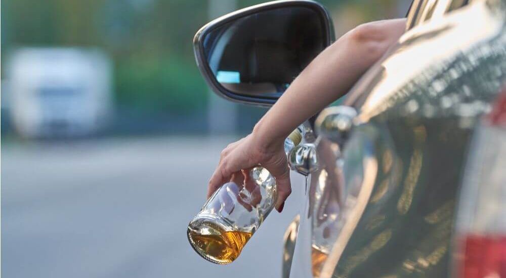 A hand holds a beer bottle outside a car window, suggesting an action of discarding or preparing to drink. The setting is a roadway with blurred vehicles in the background.