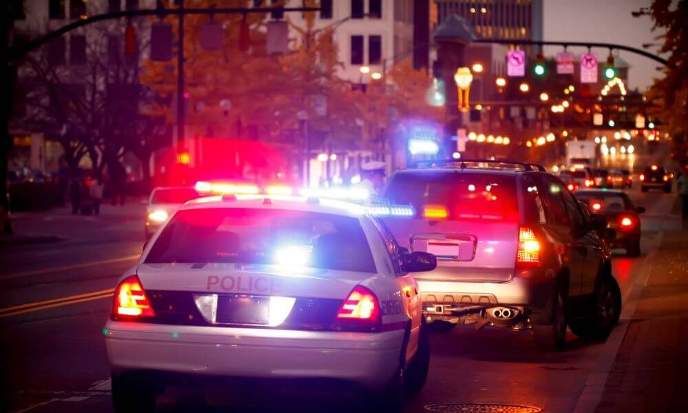 A police car, with flashing lights, is parked behind a pulled-over vehicle on a city street. Streetlights illuminate autumn trees and buildings as cars pass by in the background.