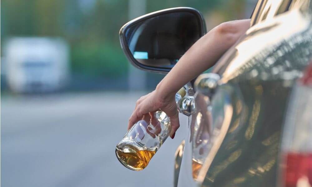 A hand holds a beer bottle outside a car window, suggesting an action of discarding or preparing to drink. The setting is a roadway with blurred vehicles in the background.
