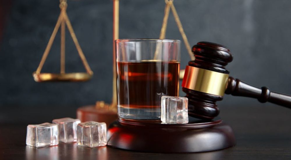 A wooden gavel rests beside a glass of dark liquid with ice cubes, while a golden balance scale looms in the background, suggesting a legal or judicial context.