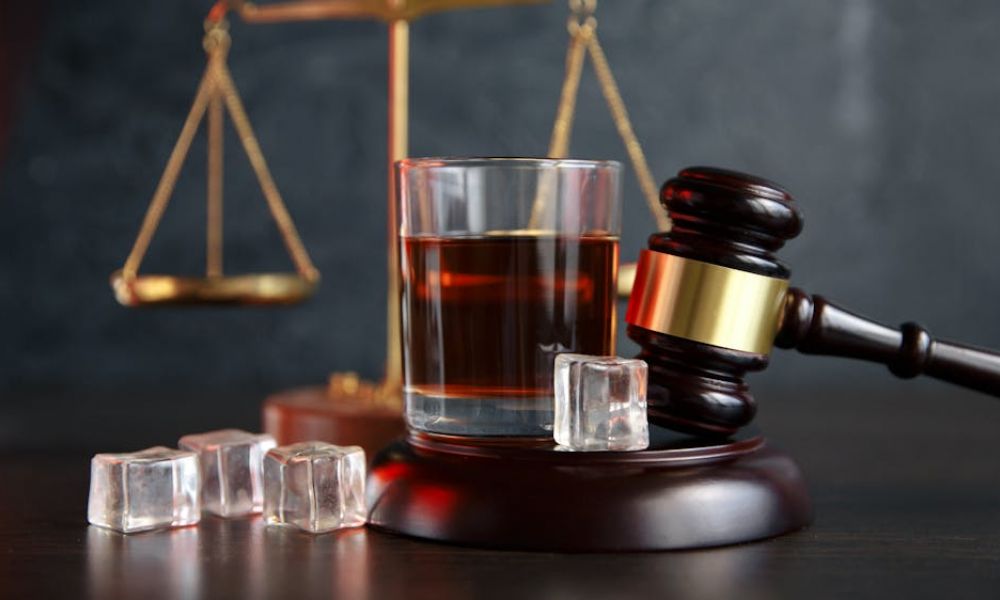 A wooden gavel rests beside a glass of dark liquid with ice cubes, while a golden balance scale looms in the background, suggesting a legal or judicial context.