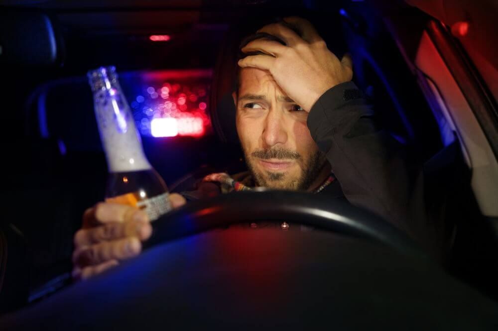 A person sits in a car holding a bottle, looking distressed. Police lights flash red and blue through the rear window, casting colored glows inside the vehicle.