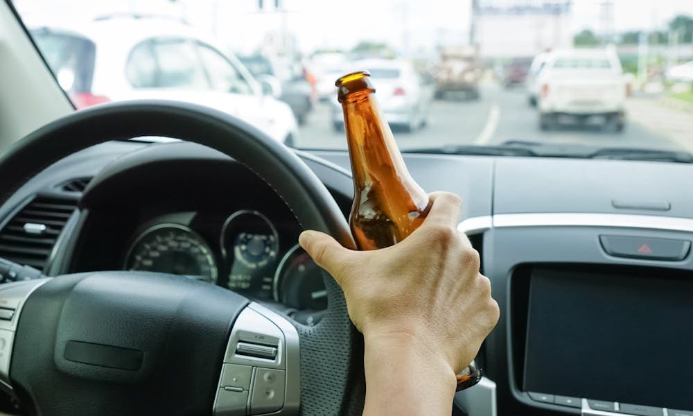 Hand holding a brown bottle peers through a car's steering wheel, dashboard visible, amidst urban traffic on a busy road.