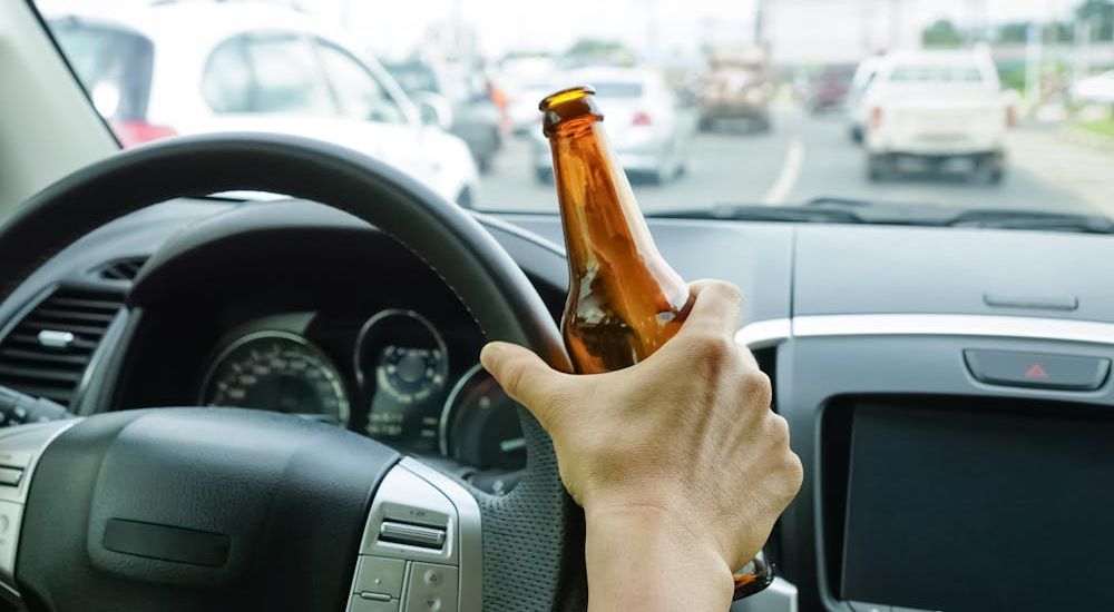 Hand holding a brown bottle peers through a car's steering wheel, dashboard visible, amidst urban traffic on a busy road.
