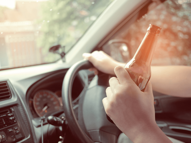 A hand holds a beer bottle while gripping the steering wheel of a car, with blurred trees and a fence visible through the windshield, suggesting a relaxed driving environment.