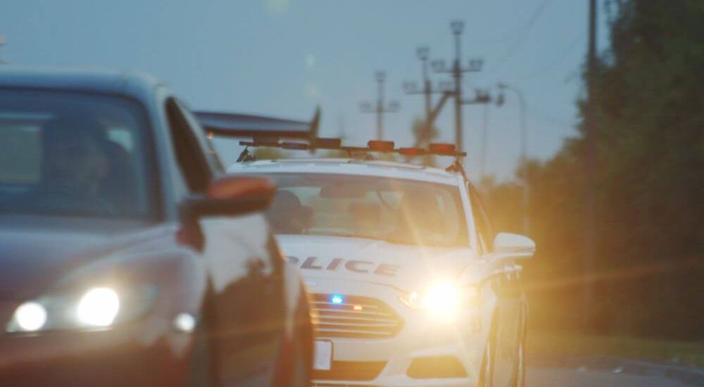 A police car with flashing lights is positioned on a road, interacting with a nearby red sedan. The environment appears dim, with power lines visible in the background.