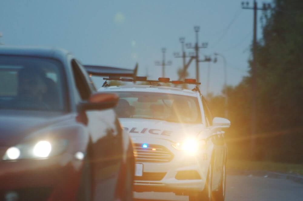 A police car with flashing lights is positioned on a road, interacting with a nearby red sedan. The environment appears dim, with power lines visible in the background.