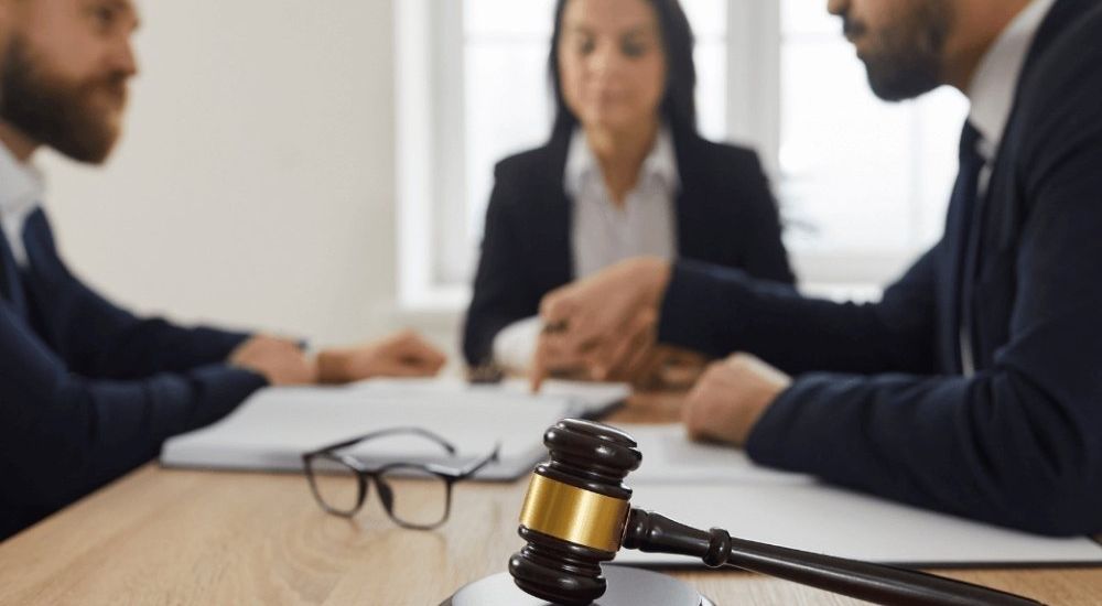 Three people in a discussion at a table with a gavel in the foreground, symbolizing a legal meeting or contract negotiation.