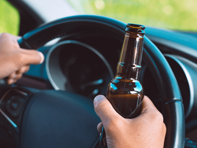 Hands gripping a steering wheel, one holding an empty brown bottle, inside a car with the dashboard partially visible, suggesting the act of drinking while driving.