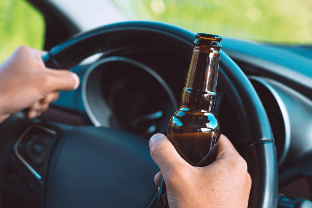 Hands gripping a steering wheel, one holding an empty brown bottle, inside a car with the dashboard partially visible, suggesting the act of drinking while driving.