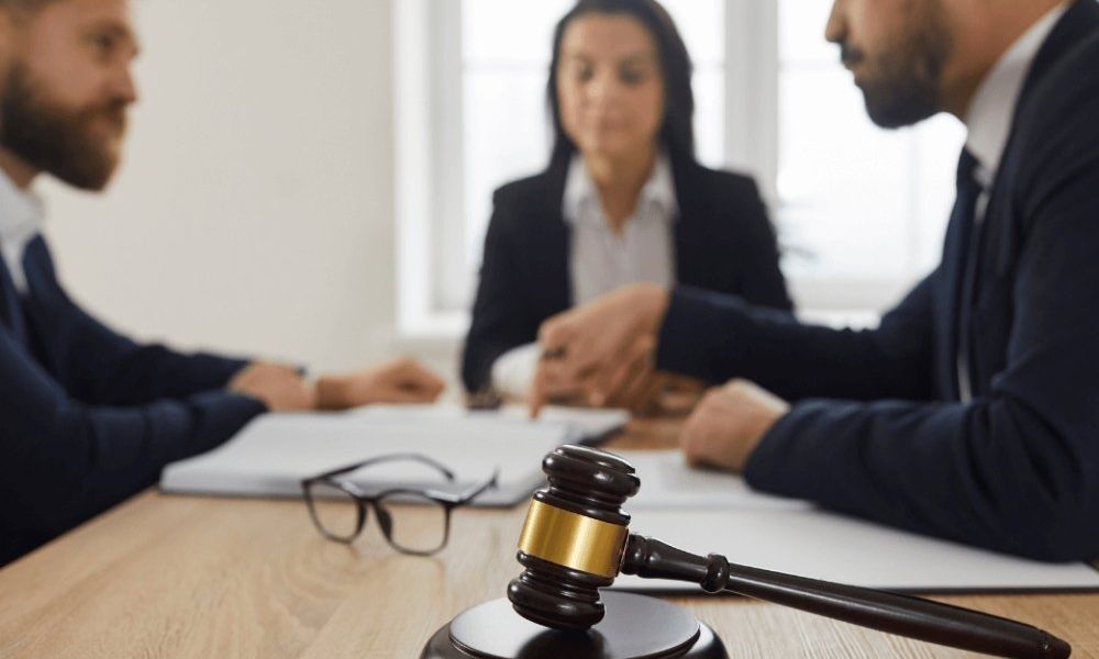 Three people in a discussion at a table with a gavel in the foreground, symbolizing a legal meeting or contract negotiation.