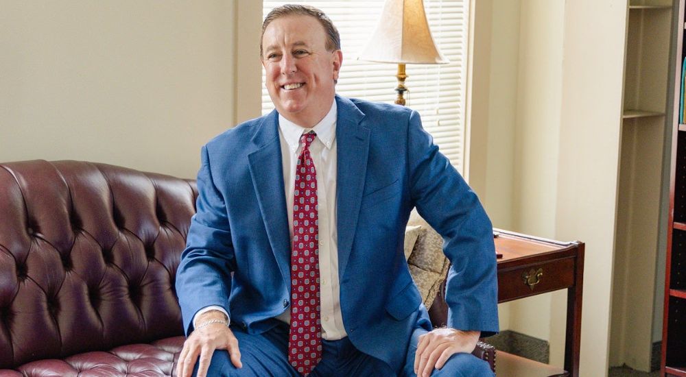 Anthony Montagna III in a blue suit and red tie sits smiling on a brown leather couch. A lamp on a side table and bookshelves are in the background.