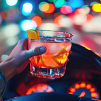 Newport-News-Lawyer-Explains-DUI-in-VA Hand holding a cocktail with lemon slice over car steering wheel, bokeh city lights in background promote drink driving risk.