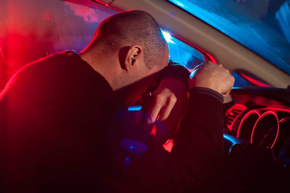 A person leans on a steering wheel, appearing distressed. Red and blue lights illuminate the car interior, suggesting a police presence outside.