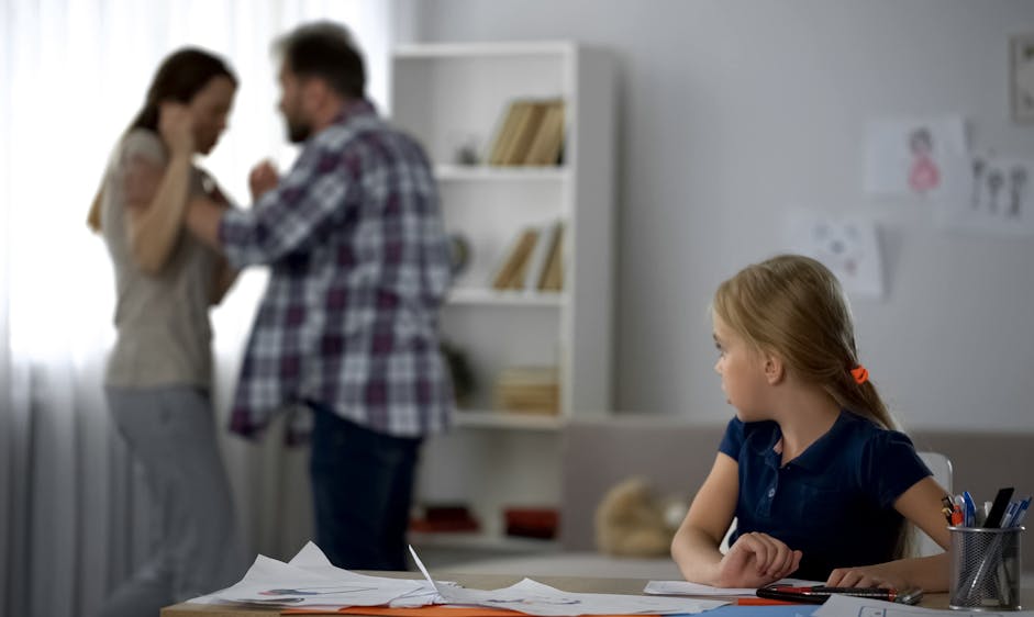 A young girl, sitting at a table with drawings, watches a couple in a tense confrontation near a bookshelf in a softly lit room. Drawings are displayed on the wall.