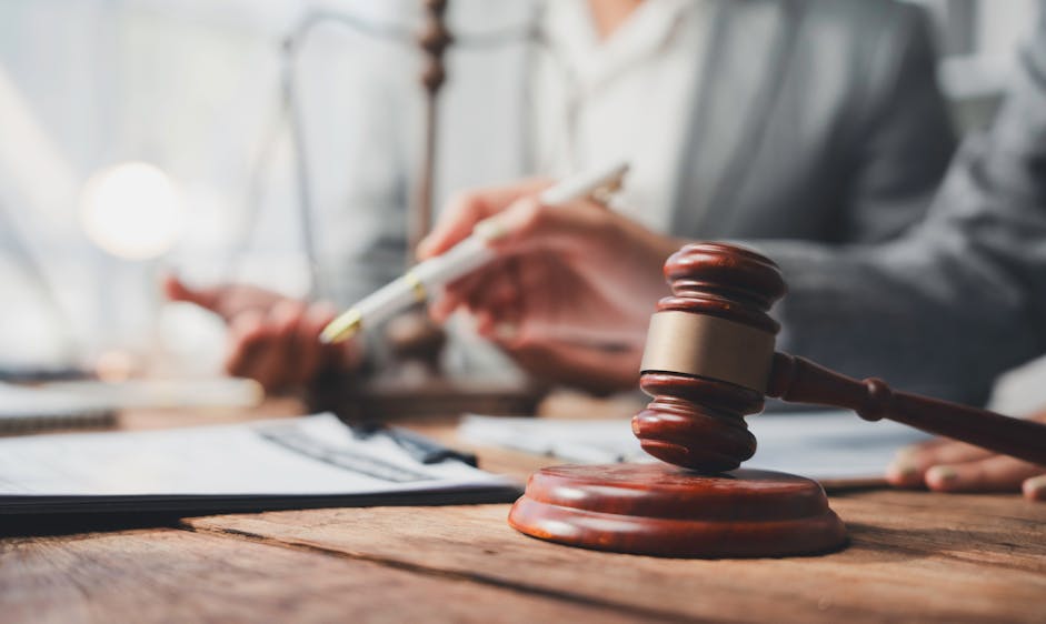 A wooden gavel rests on a table as people in suits review documents and take notes in a well-lit business setting.
