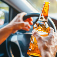 Person holding beer while driving; highlighting the dangers of drinking and driving.