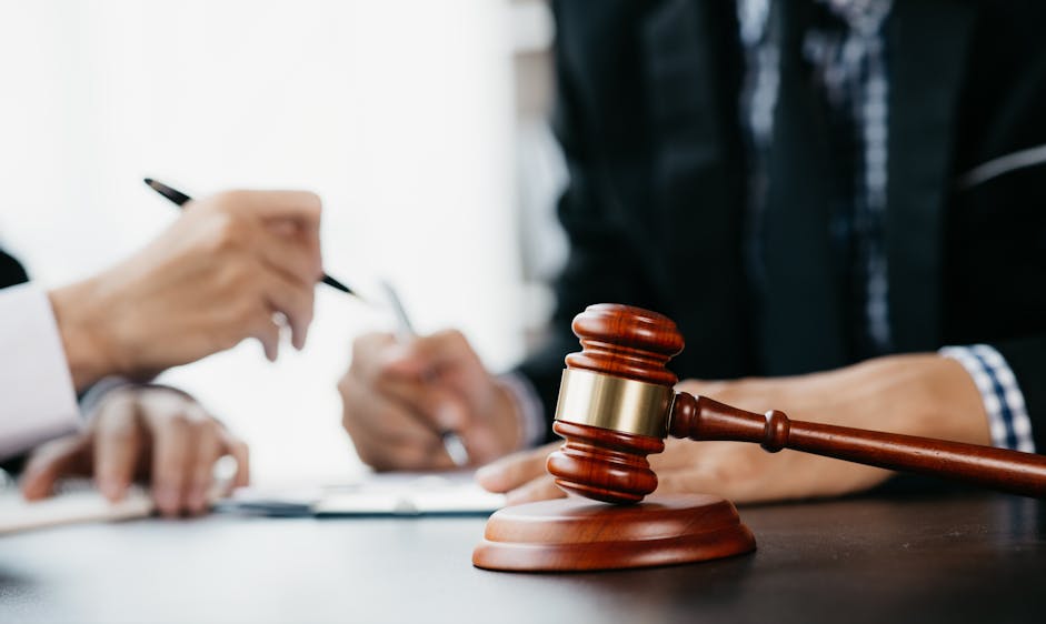 A wooden gavel rests on a table while two people in formal attire focus intently on writing notes in a bright office setting.