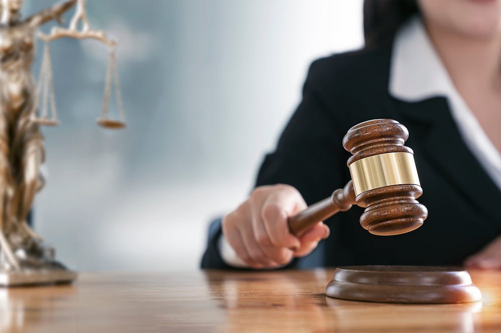 A gavel being lowered by a person in a suit onto a block in a courtroom setting, with scales of justice visible in the background.