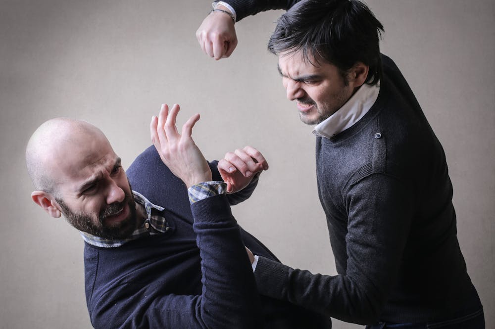 Two men engaged in a physical altercation; one is punching the other, who defensively raises his arm. They are in a neutral, indoor setting with plain background.