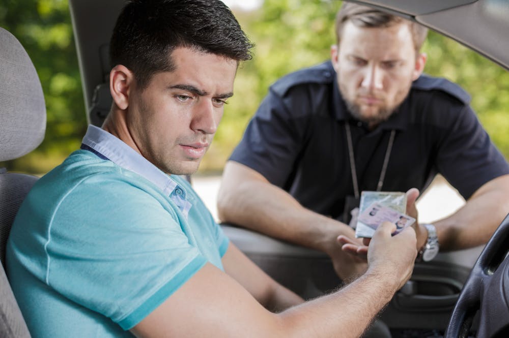 A man inside a car looks concerned while handing his identification to a police officer, who leans in from outside the vehicle. The background shows blurred greenery, indicating an outdoor setting.