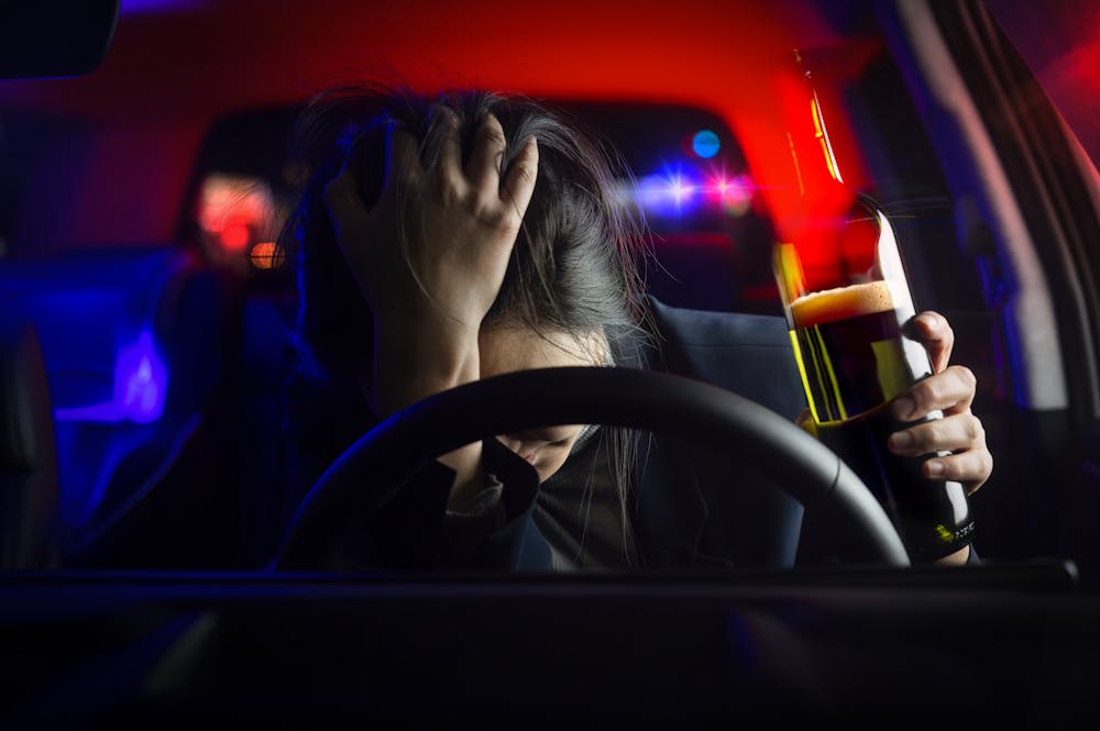 A person sits in a car, holding a bottle of alcohol with one hand while resting their head on the steering wheel, appearing distressed, amid blurred police lights in the background.
