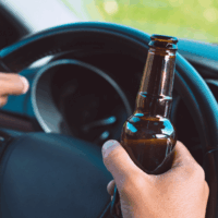 Hands gripping a steering wheel, one holding an empty brown bottle, inside a car with the dashboard partially visible, suggesting the act of drinking while driving.