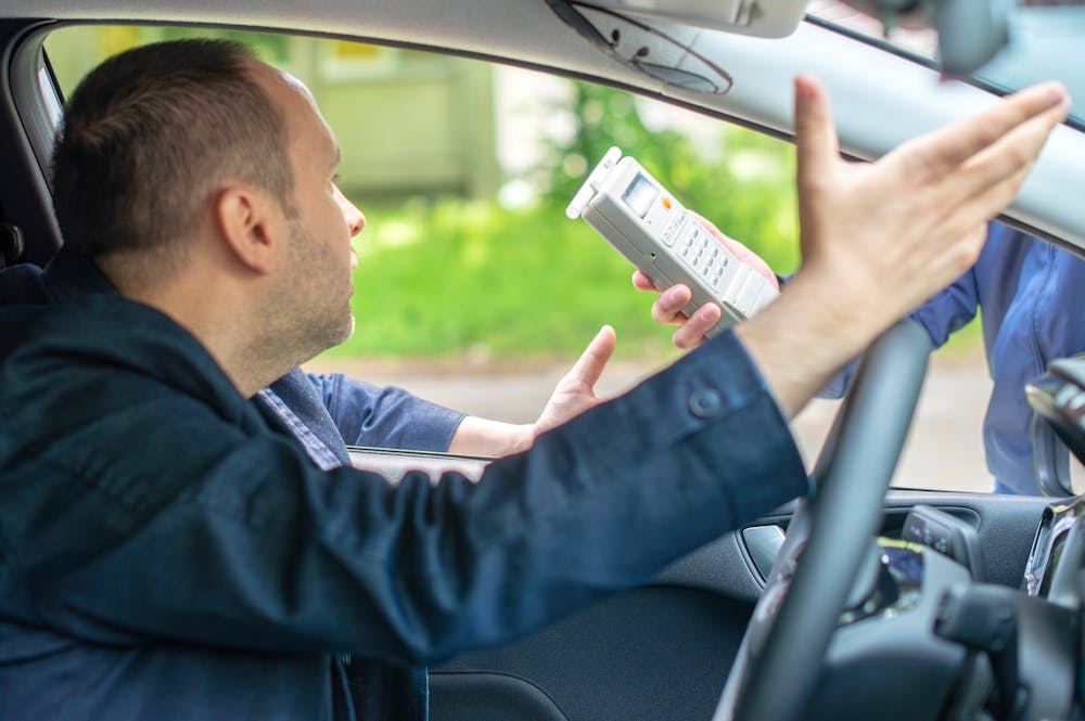 A man in a car gestures animatedly while facing another person outside, who holds a device resembling a handheld ticket printer. The scene is set in a green outdoor environment.