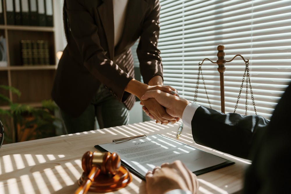 A woman shakes hands with a man at a desk, with a gavel and legal documents nearby, surrounded by a bookshelf and light filtering through blinds in an office setting.