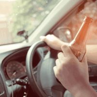 A hand holds a beer bottle while gripping the steering wheel of a car, with blurred trees and a fence visible through the windshield, suggesting a relaxed driving environment.