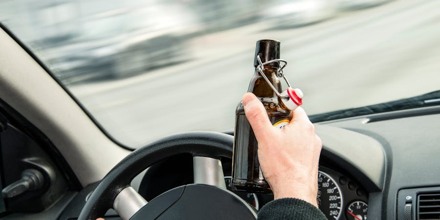 A hand grips a brown glass bottle while steering a car, indicating driving with one hand. The blurred background suggests motion, depicting a potentially unsafe situation on the road.