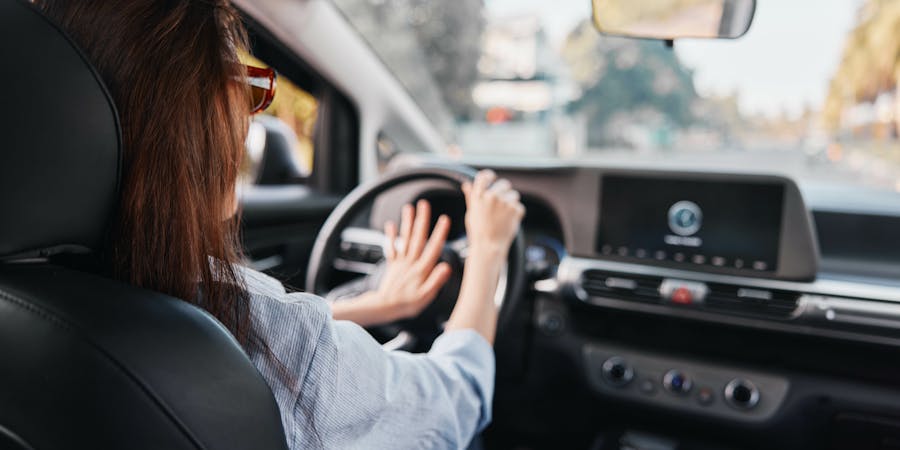 A woman is driving a car, holding the steering wheel with one hand and gesturing with the other. The interior features a modern dashboard with a digital screen, surrounded by road scenery.