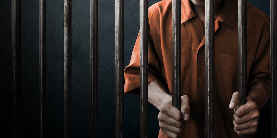 A person in an orange prison uniform grips metallic bars, suggesting confinement. The setting is dark and somber, reinforcing a sense of captivity and isolation.