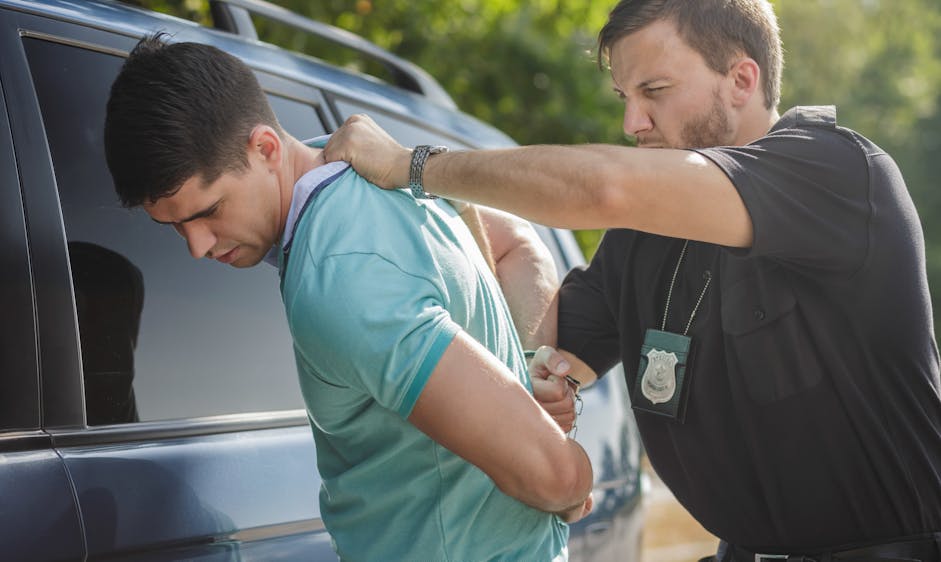 A police officer is restraining a man in a green shirt against a car, with the officer's badge visible, in a wooded, outdoor environment. The man appears apprehensive.