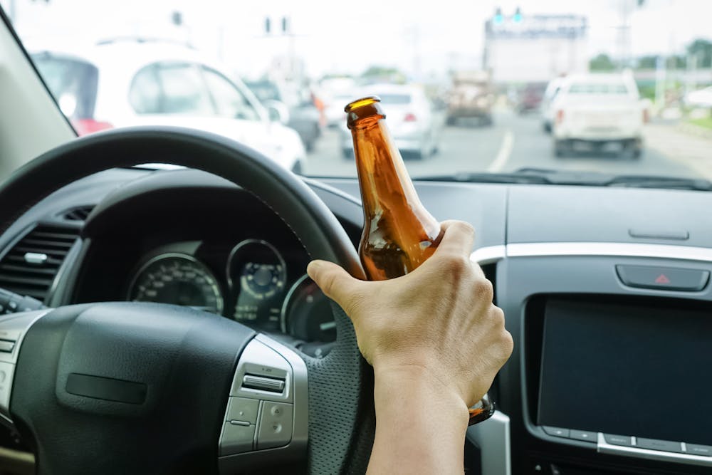 Hand holding a brown bottle peers through a car's steering wheel, dashboard visible, amidst urban traffic on a busy road.