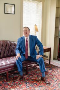 Attorney Anthony Montagna, III in blue suit sitting on a leather couch in an office with a lamp and colorful rug.