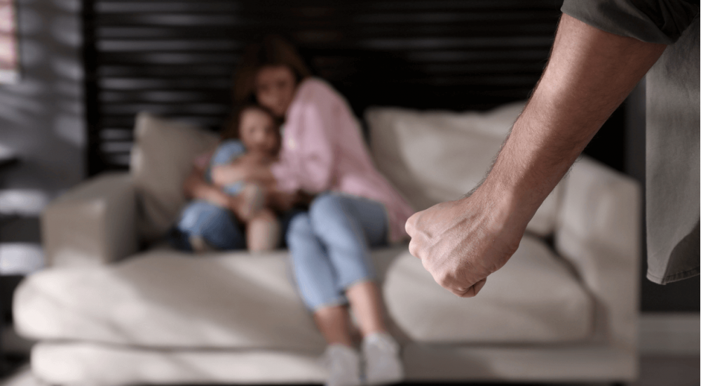 A clenched fist is prominently foregrounded, poised and tense. In the blurred background, a woman and child sit on a couch, appearing vulnerable in a room with soft lighting.