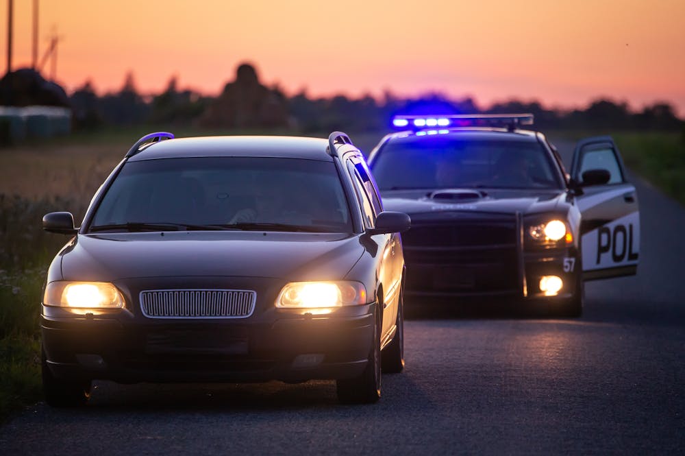 A car stopped on a rural road, with headlights on, is followed by a police car with flashing blue lights and an open door.